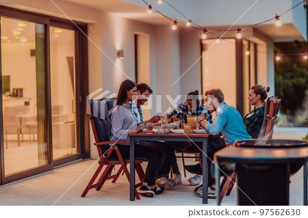 A group of young diverse people having dinner on the terrace of a modern house in the evening. Fun for friends and family. Celebration of holidays, weddings with barbecue. 97562630