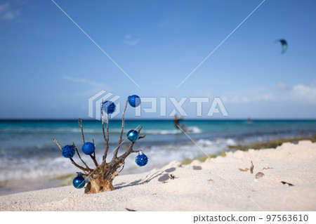 Dead corals decorated with blue Christmas balls standing on the sand beach with blue sea behind it, with a kitesurfer away.  97563610