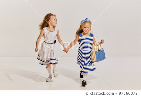 Portrait of two little girls, children in cute dresses posing, smiling, holding hands and walking over grey background Portrait of two little girls, children in cute dresses posing, smiling, holding hands and walking over grey background 97566072