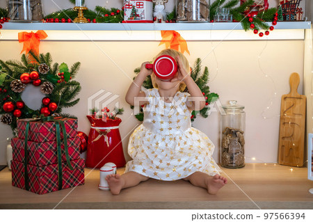 Christmas kitchen girl.Portrait of a charming little girl 2 years old, she sits on a table in a white dress and drinks from a red mug in a decorated kitchen for christmas. 97566394