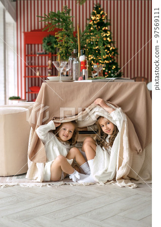 Sisters play hide-and-seek under a festive table covered with a tablecloth. Children are looking forward to the advent of Christmas Sisters play hide-and-seek under a festive table covered with a tablecloth. Children are looking forward to the advent of Christmas 97569111