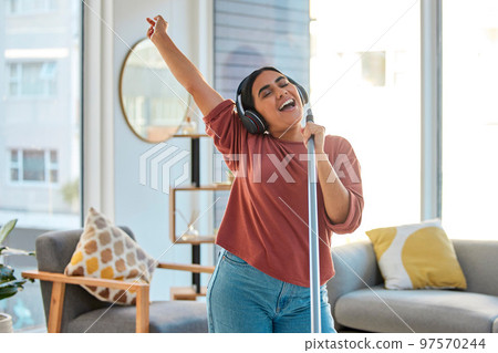 Music, headphones and woman singing while cleaning home, dancing and having fun. Singer, dance and female spring cleaning for hygiene holding broom like microphone, sweeping dust and streaming radio. 97570244