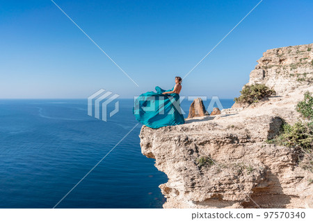A girl with loose hair in a long mint dress descends the stairs between the yellow rocks overlooking the sea. A rock can be seen in the sea. Sunny path on the sea from the rising sun 97570340