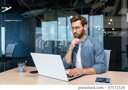 .Serious pensive businessman in shirt thinking about decision sitting at table in modern office, man with beard is using laptop at work. 97572526