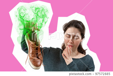 A young woman holding a sneaker in her hand, which strongly stinks. The concept of advertising insoles for shoes. Pink background. Copy space A young woman holding a sneaker in her hand, which strongly stinks. The concept of advertising insoles for shoes. Pink background. Copy space 97572885