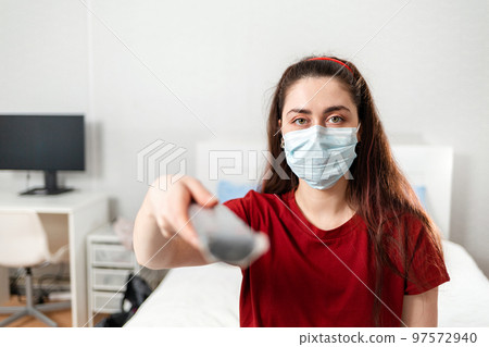 Portrait of a Young woman in a medical mask holding a TV remote control. The concept of disinformation and false news 97572940