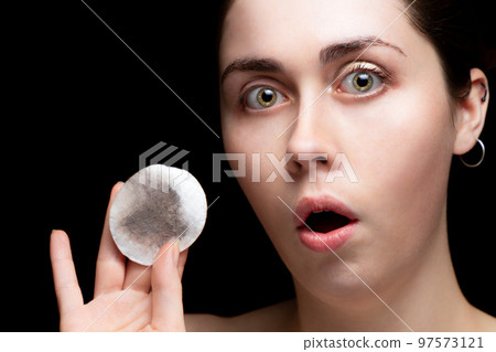 Beautiful girl surprised and shows a cotton pad with makeup. Black background 97573121
