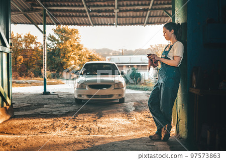 The concept of women's equality and feminism. A woman in a work uniform stands against the wall and wipes her hands with a rag. In the background, the setting sun and the car 97573863