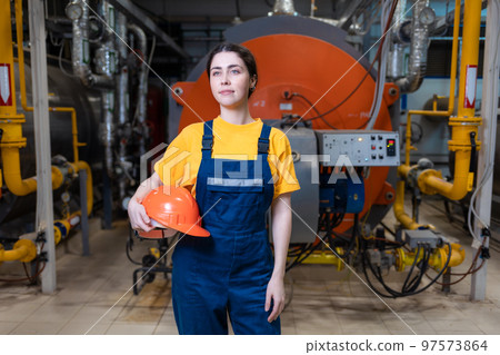 The concept of industrial manufactory. A young smiling woman in uniform holds a safety helmet in her hand and poses while standing in the boiler room 97573864