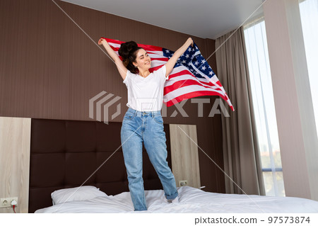 Young happy woman having fun with an American flag on her bed. Indoor. The concept of national American holidays and patriotism Young happy woman having fun with an American flag on her bed. Indoor. The concept of national American holidays and patriotism 97573874