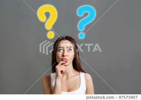 The concept of searching for ideas and information. Portrait of a brooding young Caucasian woman looking up at question marks. Gray background. Copy space The concept of searching for ideas and information. Portrait of a brooding young Caucasian woman looking up at question marks. Gray background. Copy space 97573907