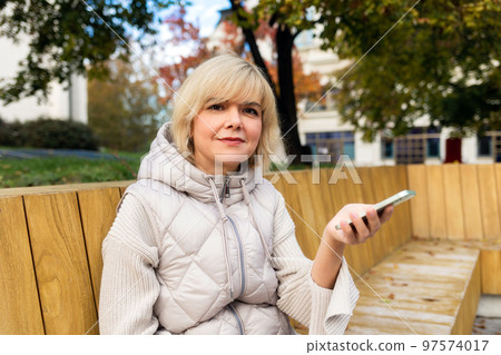 Portrait of adult smiling Caucasian woman is sitting on bench and video calls with smartphone. Concept of online freelance 97574017