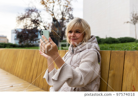 Free Wi-Fi in city park. Portrait of adult smiling Caucasian woman is sitting on bench and video calls with smartphone. Concept of online communication and social web Free Wi-Fi in city park. Portrait of adult smiling Caucasian woman is sitting on bench and video calls with smartphone. Concept of online communication and social web 97574018