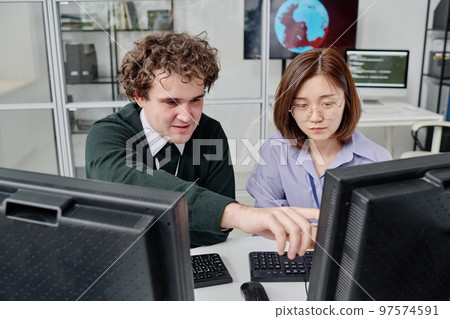 Two young colleagues working with security of computer program together, they sitting at table with computer monitors Two young colleagues working with security of computer program together, they sitting at table with computer monitors 97574591