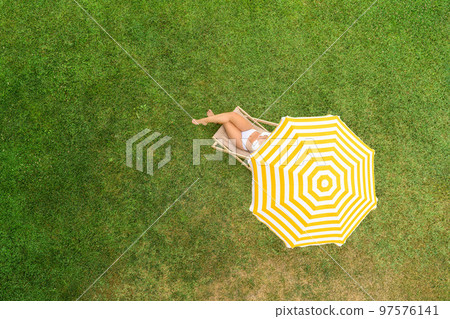 Woman in a white bikini sitting on deck chair under yellow umbrella on the green grass sunbathes at summer day. Top view, drone, aerial view, Woman in a white bikini sitting on deck chair under yellow umbrella on the green grass sunbathes at summer day. Top view, drone, aerial view, 97576141