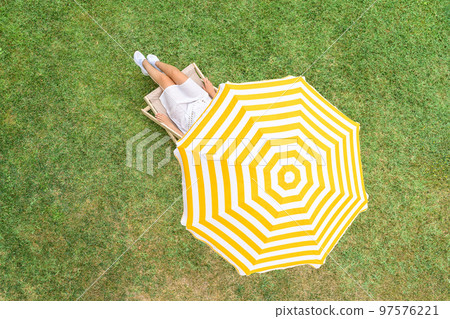 Woman in a white dress sitting on deck chair under yellow umbrella  on the green grass sunbathes at summer day. Top view, drone, aerial view. 97576221