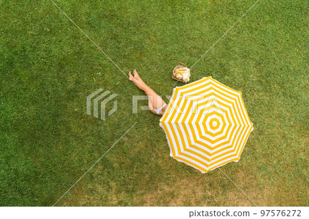 Woman in white bikini with basket for picnic lying under yellow umbrella on the green grass sunbathes at summer day. Top view, drone, aerial view. Woman in white bikini with basket for picnic lying under yellow umbrella on the green grass sunbathes at summer day. Top view, drone, aerial view. 97576272