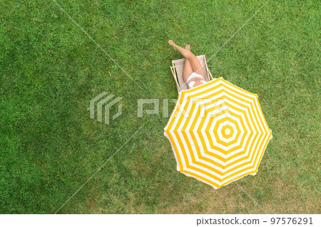Woman in a white bikini sitting on deck chair under yellow umbrella  on the green grass sunbathes at summer day. Top view, drone, aerial view. 97576291