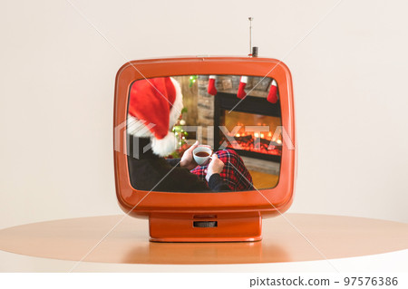 Tv showing movie of One divorce man with cup coffee, tea in santa claus hat sitting and warming at winter evening near fireplace flame, covered christmas plaid 97576386