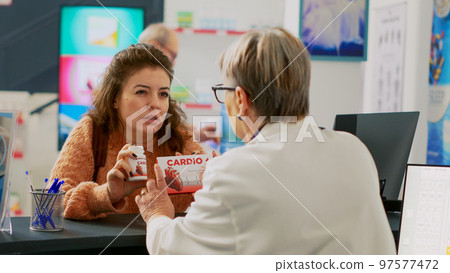 Woman showing cardiology pills to pharmacist, asking about medical support to buy treatment. Person looking at boxes of supplements and drugs to buy at pharmacy cash register. Handheld shot. Woman showing cardiology pills to pharmacist, asking about medical support to buy treatment. Person looking at boxes of supplements and drugs to buy at pharmacy cash register. Handheld shot. 97577472