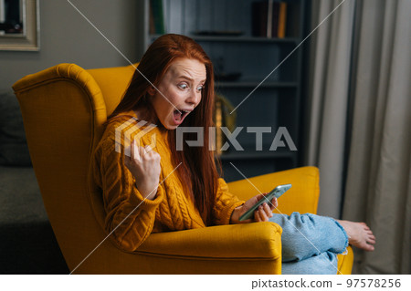 Portrait of happy excited young woman looking mobile phone screen sitting in yellow armchair, laughing with good news, feeling great positive surprise reaction receiving gift reading sms at home. Portrait of happy excited young woman looking mobile phone screen sitting in yellow armchair, laughing with good news, feeling great positive surprise reaction receiving gift reading sms at home. 97578256