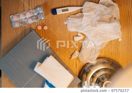 High-angle close-up view of home sickness medical supplies lying on bedside table beside the bed. Top closeup view of electronic thermometer, fever pills, warming ointment next to lamp. High-angle close-up view of home sickness medical supplies lying on bedside table beside the bed. Top closeup view of electronic thermometer, fever pills, warming ointment next to lamp. 97578257