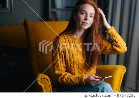 Portrait of gorgeous redhead young woman using smartphone sitting in yellow armchair, smiling looking at camera. Attractive ginger female holding phone in hands sitting in soft cozy chair at home 97578285