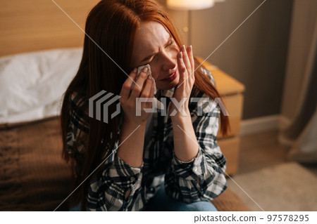 Close-up portrait of unhappy depressed redhead woman crying and wiping tears with paper tissue, suffer from grief, divorce or break up with boyfriend, broken heart, sitting on bed at home. 97578295