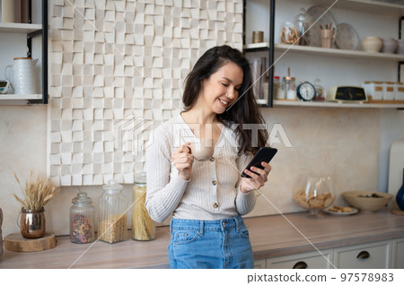Happy young woman using smartphone while drinking morning tea or coffee in kitchen interior at home, free space Happy young woman using smartphone while drinking morning tea or coffee in kitchen interior at home, free space 97578983