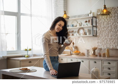 Happy caucasian woman using laptop and drinking tea, sitting on table in kitchen and surfing internet, free space 97578985