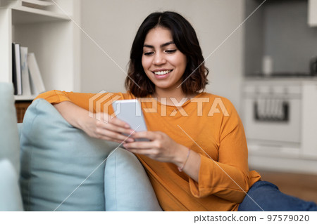 Smiling Arab Woman Using Smartphone While Relaxing On Couch At Home 97579020
