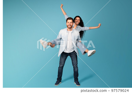 Cheerful young european guy hold woman in casual on back, have fun, isolated on blue background, studio 97579024