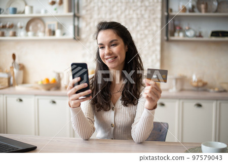 Happy caucasian woman buying food online, using cellphone and plastic credit card, shopping online, sitting in kitchen 97579043