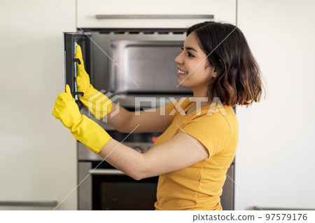 Housekeeping Concept. Smiling Arab Woman In Rubber Gloves Wiping Microwave From Dirt Housekeeping Concept. Smiling Arab Woman In Rubber Gloves Wiping Microwave From Dirt 97579176