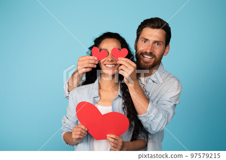 Glad young european husband in casual clothes puts hearts on arab wife eyes, have fun, isolated on blue background Glad young european husband in casual clothes puts hearts on arab wife eyes, have fun, isolated on blue background 97579215