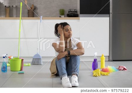 Upset tired millennial african american housewife in apron sits on floor with cleaning supplies, looks at mess 97579222