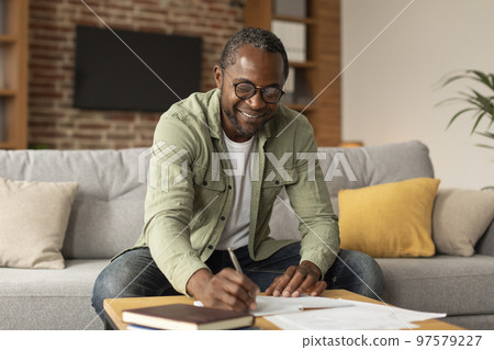 Cheerful adult black businessman in glasses makes notes, studies and works in living room interior, copy space Cheerful adult black businessman in glasses makes notes, studies and works in living room interior, copy space 97579227