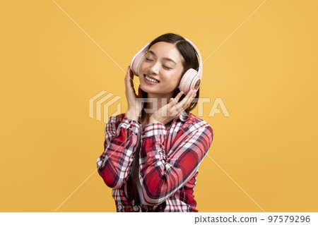 Peaceful asian woman listening to music online in wireless headphones and touching new headset, yellow background 97579296