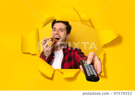 Man watching TV, pointing remote controller and eating pizza, posing in hole of torn yellow paper background Man watching TV, pointing remote controller and eating pizza, posing in hole of torn yellow paper background 97579353