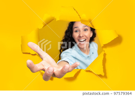 Excited woman stretching open palm through hole in torn yellow paper, taking something, holding invisible object 97579364