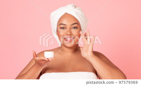 Beautiful african american oversize woman applying facial cream, holding jar in hand and smiling, pink background 97579369