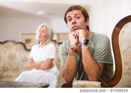 Upset man sitting while quarrel with wife at home interior 97580024