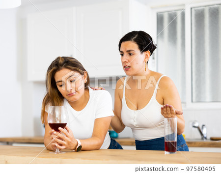 Upset woman drinking wine in kitchen while girlfriend consoling her Upset woman drinking wine in kitchen while girlfriend consoling her 97580405