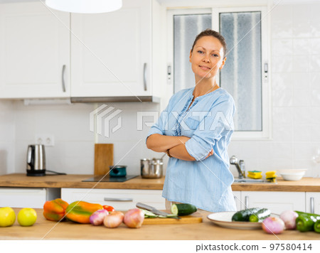 Positive housewife leaning on a kitchen set with vegetables while preparing salad Positive housewife leaning on a kitchen set with vegetables while preparing salad 97580414