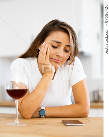Frustrated young Hispanic woman with phone drinking wine in home kitchen 97580426