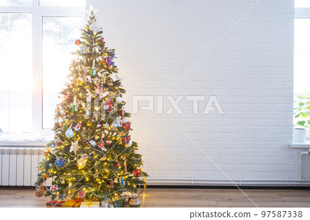 Christmas tree in the white interior of a house with large windows. Glowing fairy lights garlands interior decoration of the studio room. Potted plants in the home Christmas tree in the white interior of a house with large windows. Glowing fairy lights garlands interior decoration of the studio room. Potted plants in the home 97587338