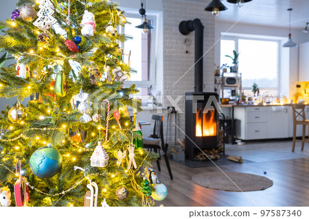 Festive interior of house is decorated for Christmas and New Year in loft style with black stove, fireplace, Christmas tree. Warm studio room with set table, burning wood, cozy and heating of home 97587340