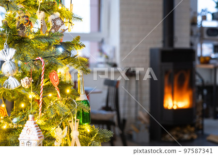 Festive interior of house is decorated for Christmas and New Year in loft style with black stove, fireplace, Christmas tree. Warm studio room with set table, burning wood, cozy and heating of home 97587341
