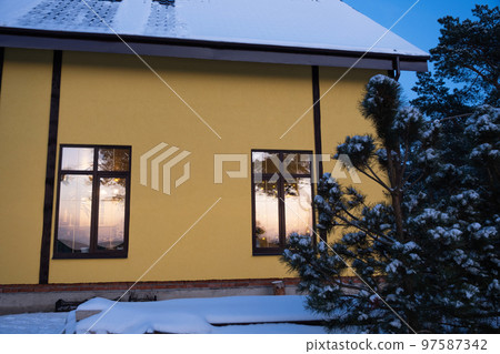 Windows of the yellow house are outside in the snow in winter. Inside the dwelling there is a warm light, fairy lights of garlands, a Christmas tree. Cozy celebration of Christmas and New Year at home Windows of the yellow house are outside in the snow in winter. Inside the dwelling there is a warm light, fairy lights of garlands, a Christmas tree. Cozy celebration of Christmas and New Year at home 97587342