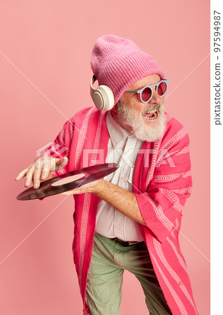 Emotive senior, old man in stylish bright outfit with knitted hat, sunglasses, headphones and vinyl over pink background. Dj Emotive senior, old man in stylish bright outfit with knitted hat, sunglasses, headphones and vinyl over pink background. Dj 97594987